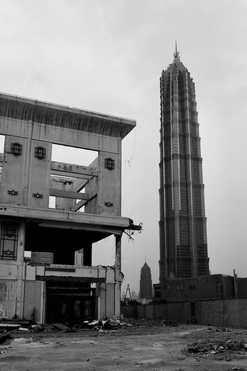 On the left, a partially demolished building reveals empty openings and hanging cables. On the right, a tall multi-storey tower stands out behind a plot covered in rubble.