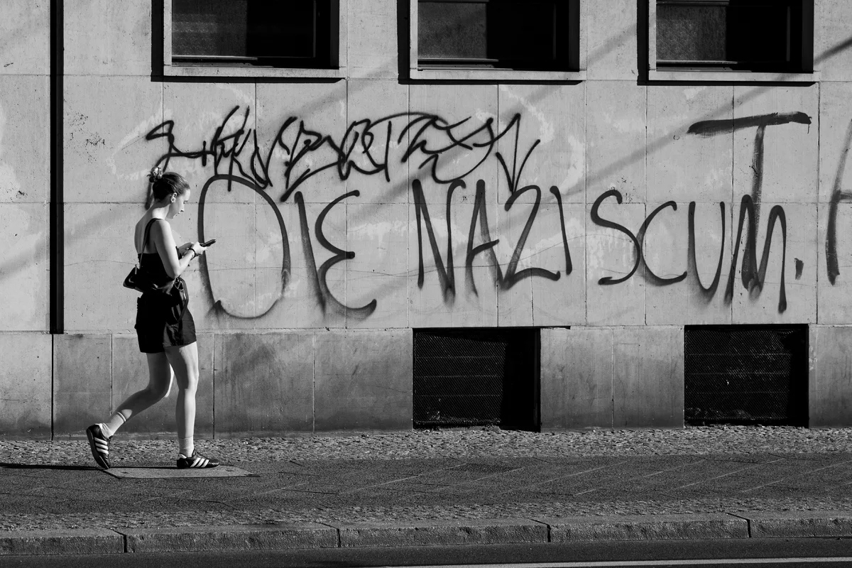 A woman walks along a pavement while looking at her phone. Behind her, a graffiti-covered wall has three windows at the top and two ventilation grilles near the ground.