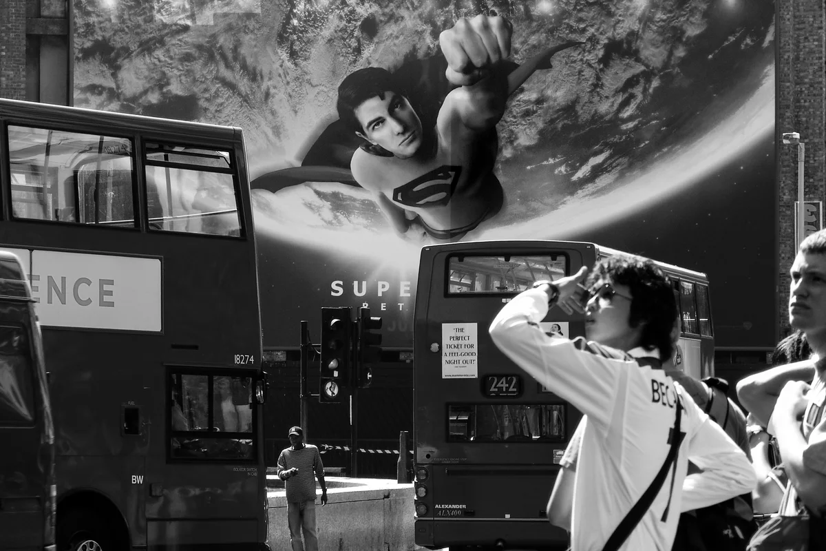 Two double-decker buses fill the street in front of a large Superman poster. Passers-by stand in the foreground, including a person in a white vest raising their hand to their face.