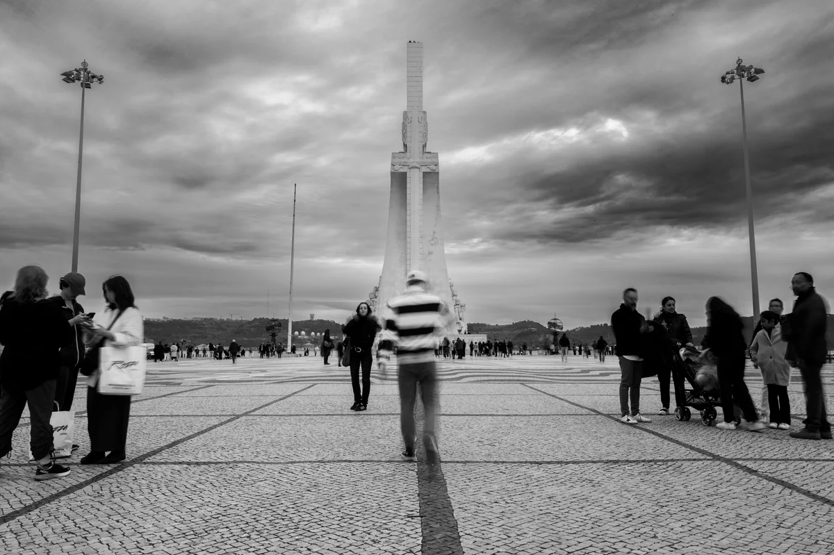 Un monument haut se dresse au centre d’une vaste place pavée, sous un ciel nuageux. Des groupes de personnes se tiennent de chaque côté, tandis qu’une silhouette floue marche au premier plan.